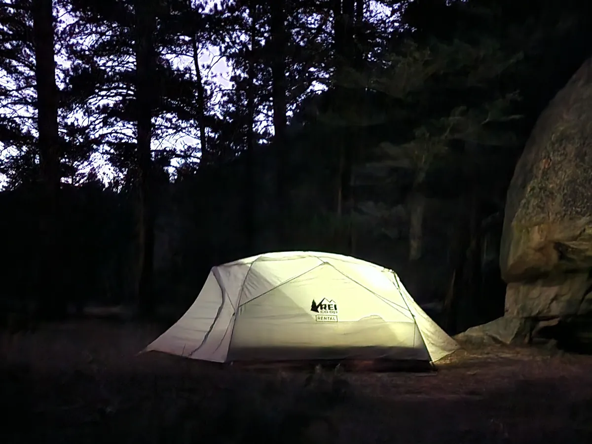 A tent glowing from inside at night, pitched beside a huge boulder under tall pines.
